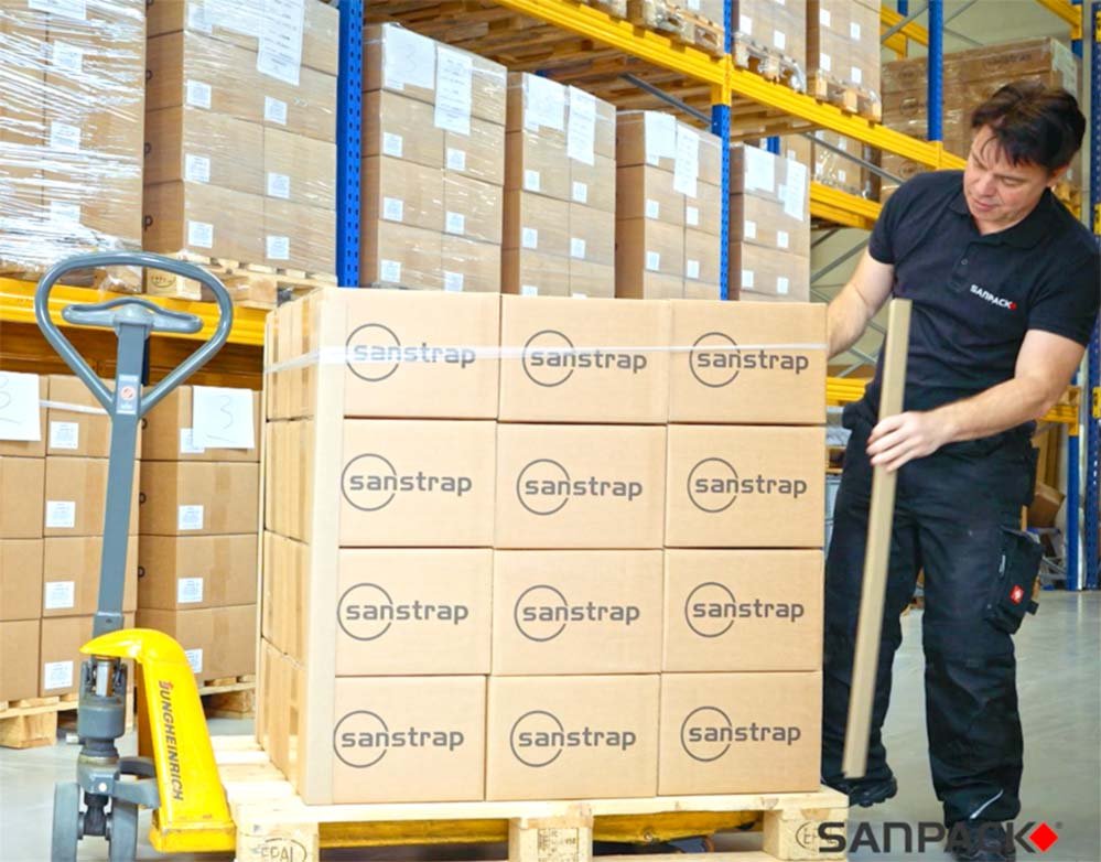 Ein Mitarbeiter sichert einen hohen Kartonstapel im Lager mit sanstrap® Dehnbändern auf einer Palette.